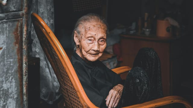 Portrait of an elderly woman sitting in a rocking chair indoors with a contemplative look.