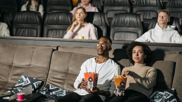 A couple enjoys popcorn while watching a movie in a cozy cinema environment, surrounded by others.