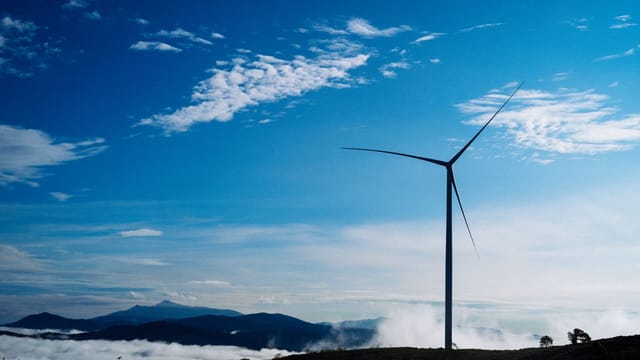A solitary wind turbine rises against a vibrant blue sky, surrounded by clouds and distant mountains.