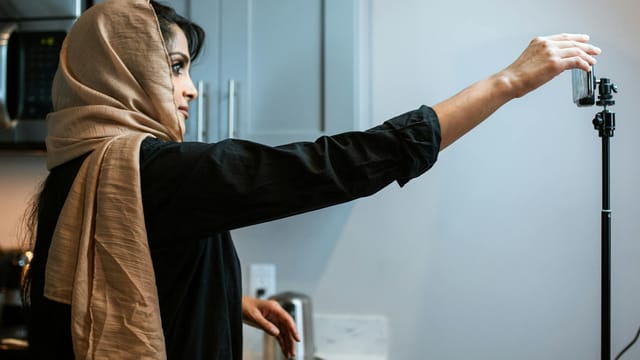 A Muslim woman in a hijab records a cooking video in her kitchen, focusing on her food presentation.
