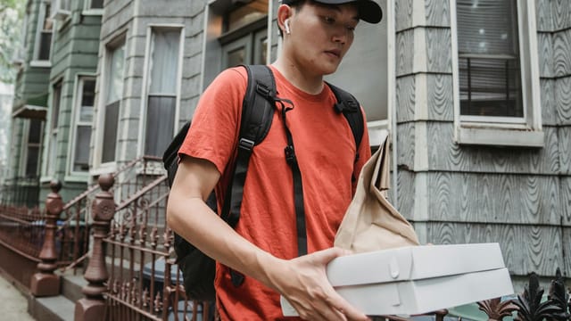 Courier delivering food in an urban neighborhood, holding pizza boxes and a brown paper bag.
