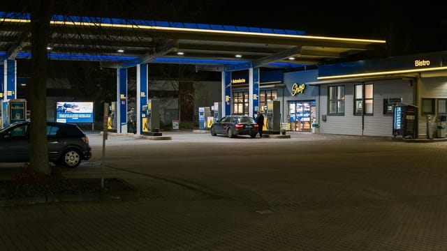 A well-lit gas station at night featuring cars and a bistro, creating a lively urban scene.