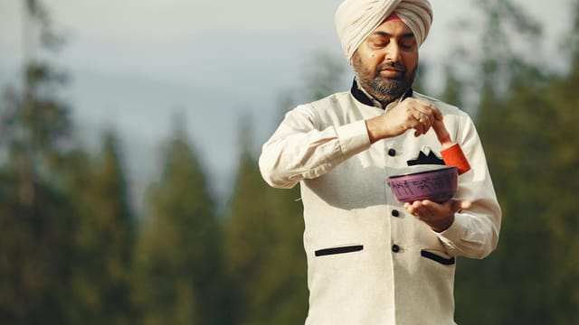 A man in traditional attire using a Tibetan singing bowl outdoors, surrounded by nature.