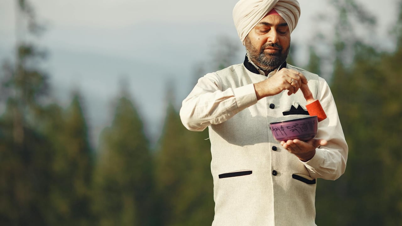 A man in traditional attire using a Tibetan singing bowl outdoors, surrounded by nature.