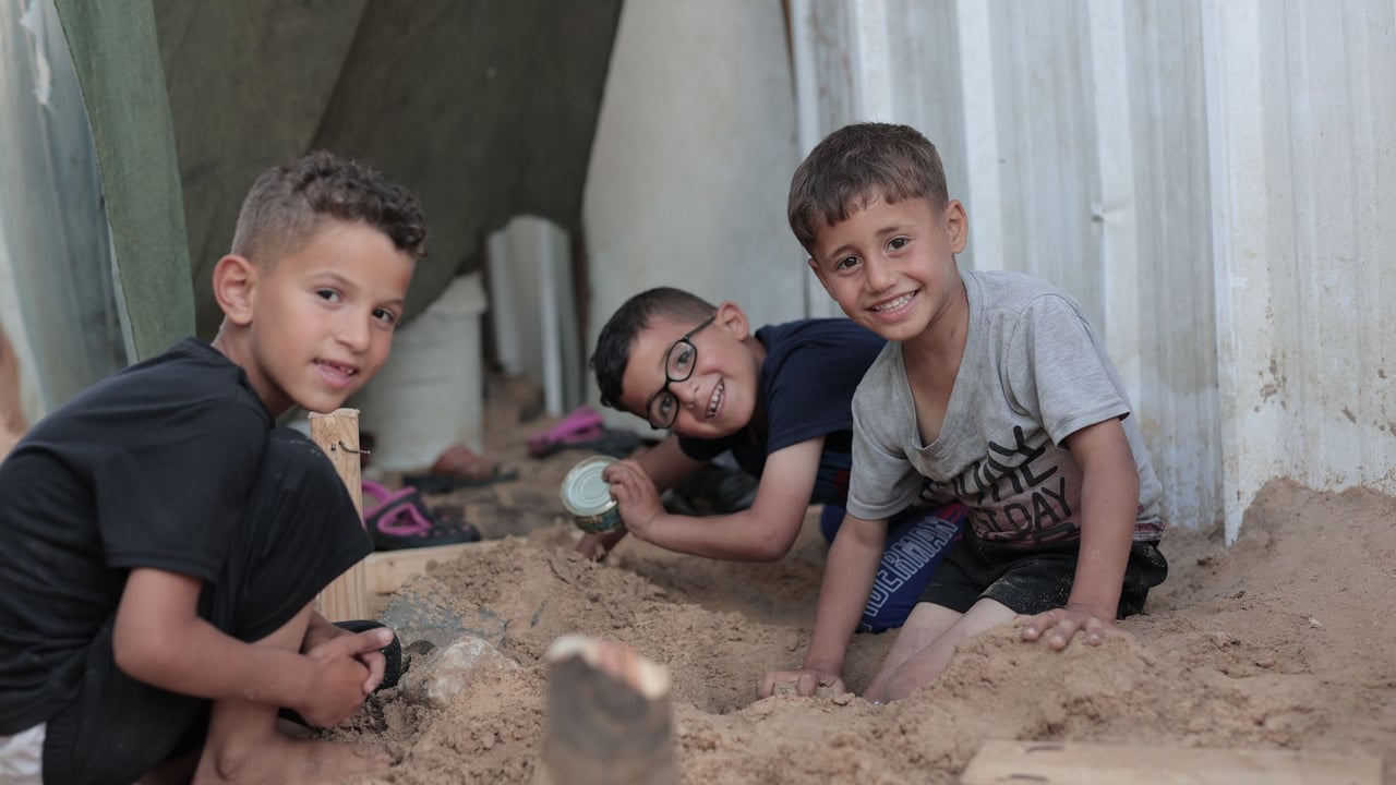 Three boys joyfully play together in the sand outdoors in Gaza, highlighting childhood innocence and friendship.