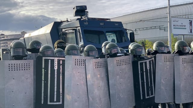Riot police in protective gear and shields in Minsk, standing in front of an armored vehicle.