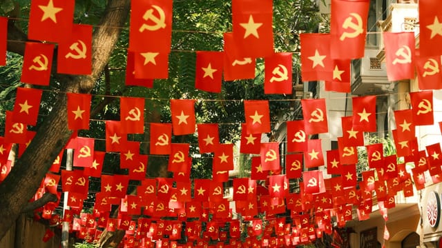 Red banners with communist symbols hang above a lively street scene, creating a vibrant display.