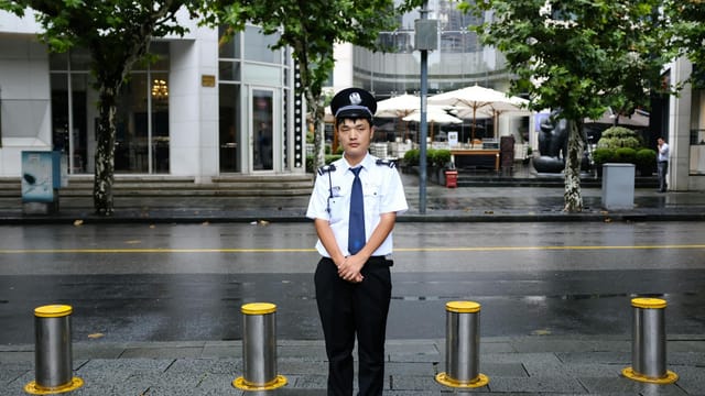 A security officer stands on a wet street in Shanghai after rain, with urban greenery and buildings.