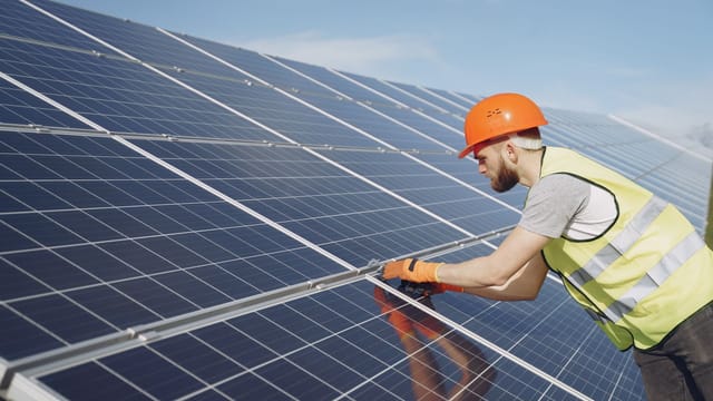 Side view of focused young man in uniform and hardhat checking setup of solar panels