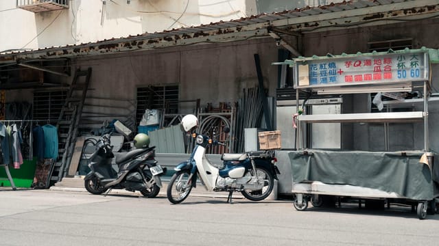 Scooters parked by a street vendor cart in Taipei, under harsh sunlight, rustic urban scene.