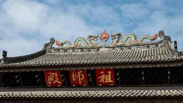 Ornate dragon sculpture atop an ancient Chinese temple roof with red banners, under a clear blue sky.