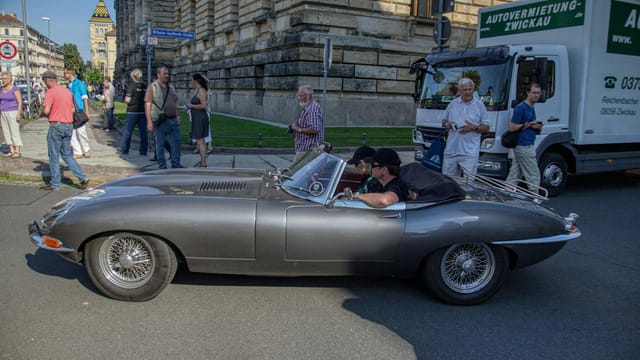 A classic Jaguar E-Type parked on a Leipzig street with people observing. Captures timeless elegance.