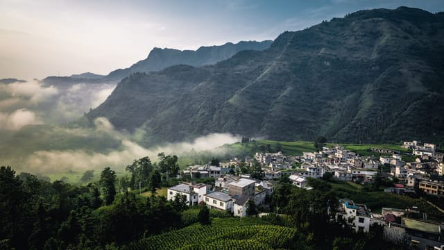 A serene village nestled in lush green mountains with morning mist drifting over the landscape.
