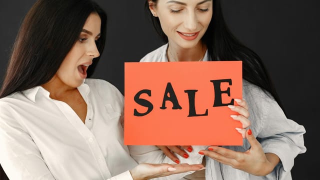 Two women energetically holding a 'SALE' sign, showcasing excitement and enthusiasm for a shopping event.
