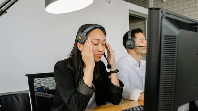 Asian woman at call center desk looking stressed, eyes closed, in office setting.