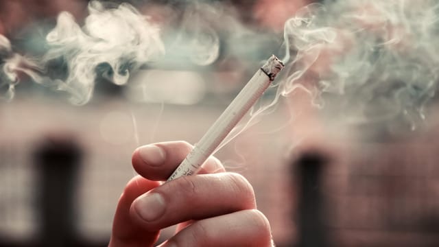 A close-up view of a hand holding a lit cigarette, with smoke visible in the air.