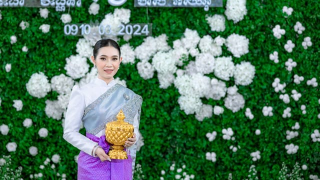 A young woman in traditional attire holding a gold urn against a floral backdrop.