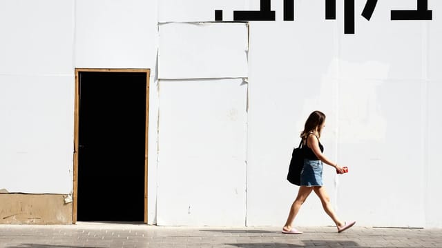 A woman walks by a white wall with Hebrew text in an urban street setting.