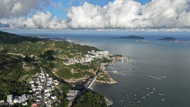 Stunning aerial view of a coastal town with lush hills and dramatic clouds overhead.