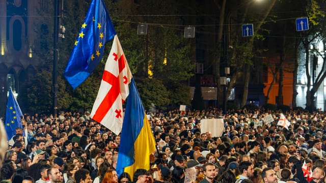 Crowd of protesters in Tbilisi holding Georgian, EU, and Ukrainian flags at night.