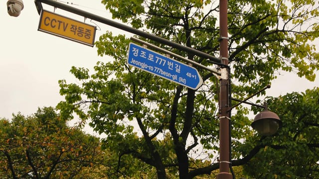 Street sign at Jeongjo-ro, Suwon, with CCTV security camera and lush trees.