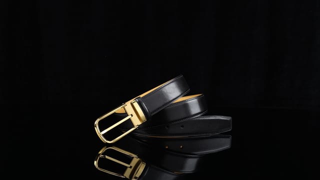 Sophisticated black leather belt with a sleek gold buckle on a reflective surface.