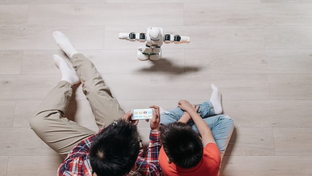 A father and son using a smartphone to control a small humanoid robot on wooden flooring.