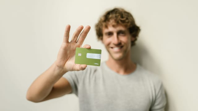 A cheerful man holding a green credit card against a neutral background.