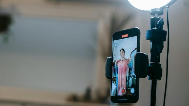 Anonymous female blogger standing in room and shooting vlog on smartphone with ring lamp while demonstrating pink dress in hands