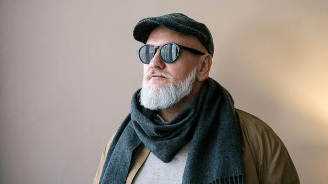 Portrait of a stylish elderly man wearing sunglasses, hat, and scarf in an indoor setting.