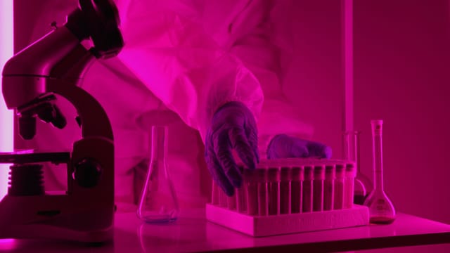 Illustrative image: Scientist in a lab coat handling test tubes under pink lighting, using a microscope.