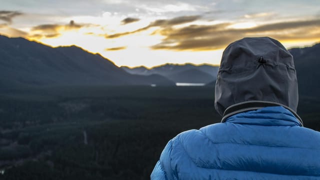 A person in a blue jacket observes a serene mountain landscape at sunset, offering a moment of tranquility.