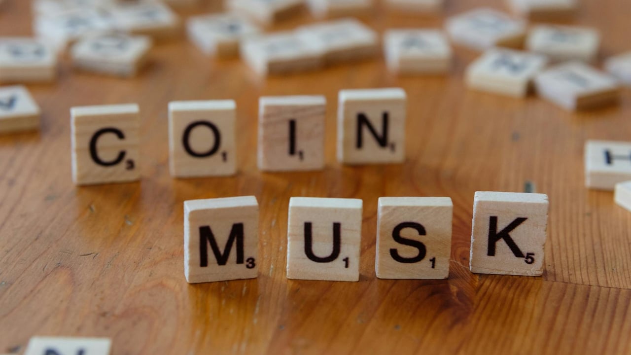 Scrabble tiles forming the words 'COIN' and 'MUSK' on a wooden table surface.