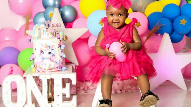 A joyful first birthday party setup with a cute child in a vibrant pink dress surrounded by balloons and a unicorn cake.