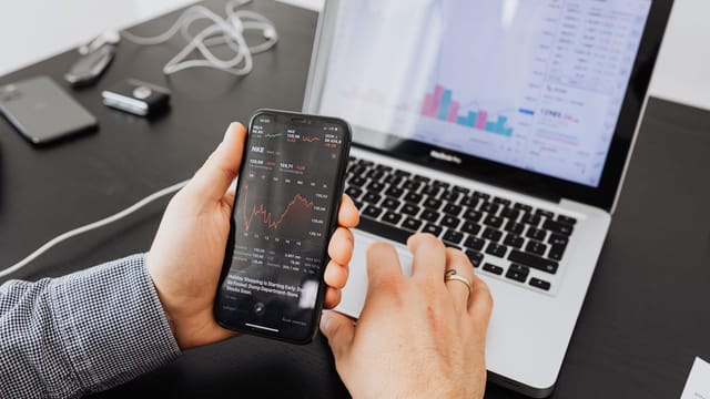Person analyzes stock data with a smartphone and laptop, indoors.