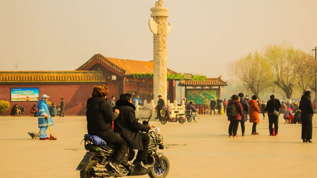 A bustling day in a Beijing square with people, scooters, and a historic Huabiao landmark.