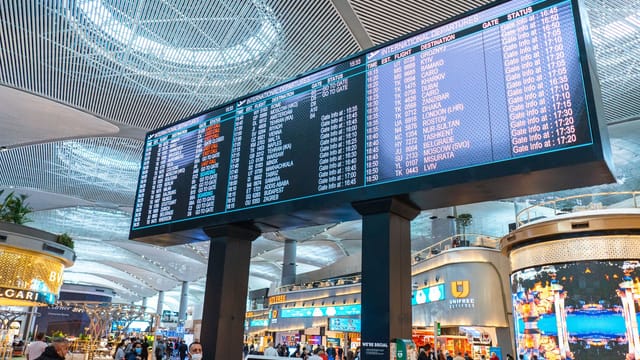 Passengers walk through Istanbul Airport's departure area, surrounded by stores and flight information displays.