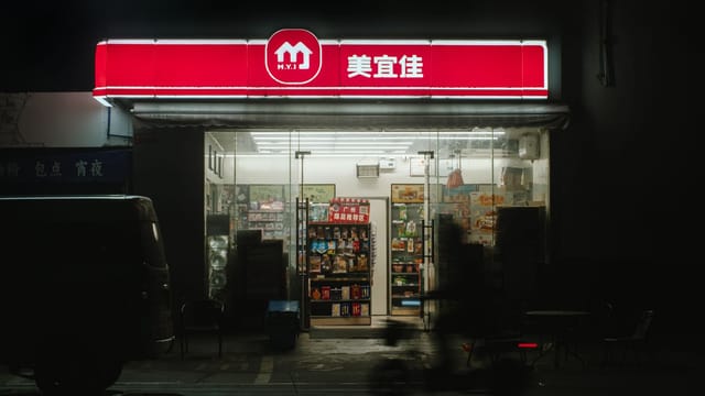 A dimly lit convenience store with a bright neon sign at night, featuring outdoor seating.