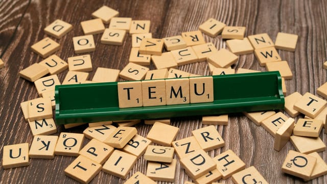 Wooden Scrabble tiles spelling TEMU on a green rack with scattered tiles on a wooden surface.