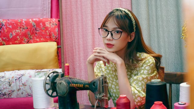 A young woman with eyeglasses sits at a sewing machine, surrounded by colorful fabrics in a textile shop.