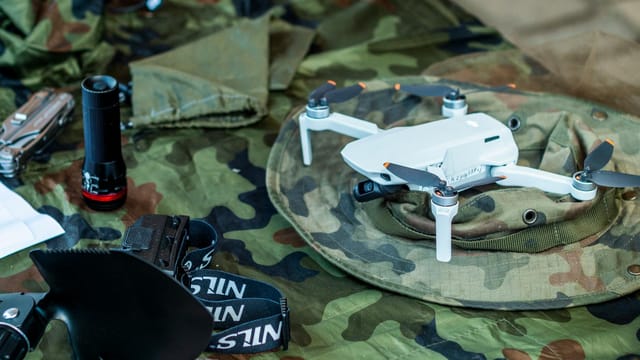 Compact white drone on camouflage gear, surrounded by flashlight and tools.