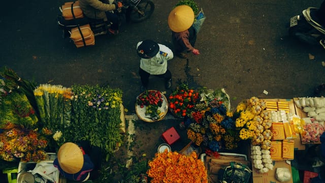 A bustling street market scene with people and colorful flowers viewed from above.