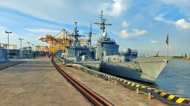 Navy vessels docked at a bustling city port with cranes and clear blue skies.