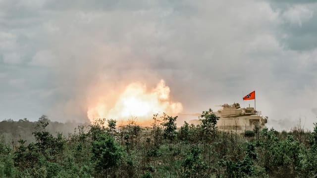 A powerful military tank firing in the woods at Fort Benning under a dramatic sky.