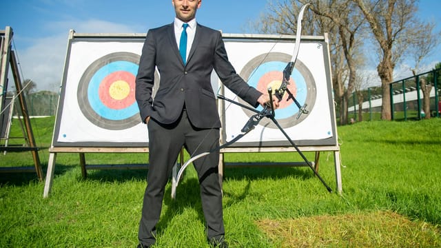 A business professional in a suit practicing archery with targets outdoors on a sunny day.