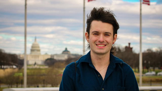 A young man smiling in front of the US Capitol in Washington, DC, with flags in the background.