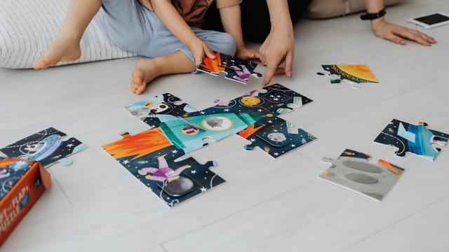 A child and adult hands putting together a colorful space-themed puzzle on the floor.