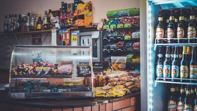 A well-stocked convenience store counter with snacks, drinks, and a refrigerator showcasing beverages.