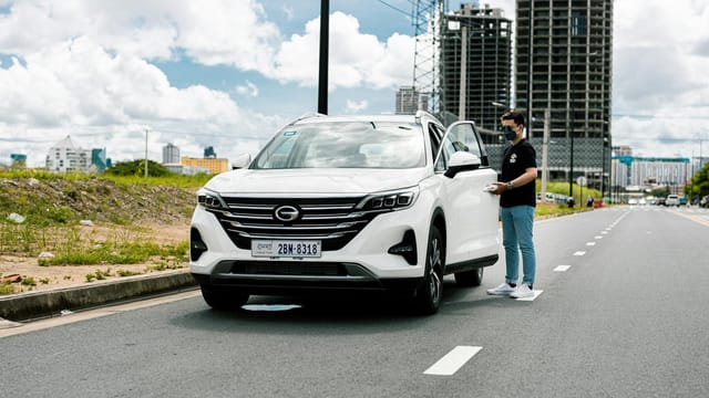 A man in a face mask stands by a parked SUV on a city road with unfinished buildings in the background.