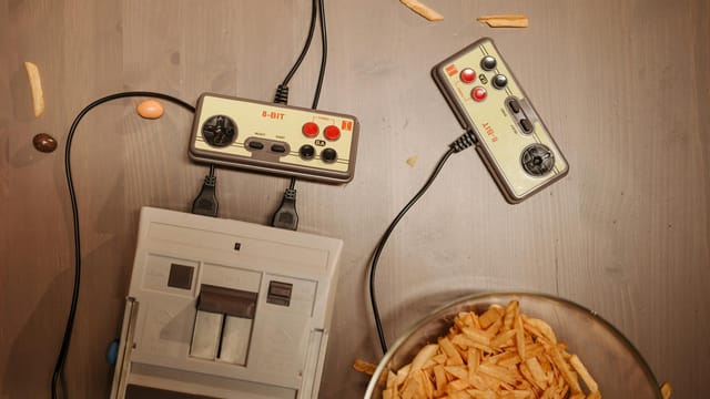 Top view of a retro gaming setup with controllers, console, and snacks on a wooden surface.